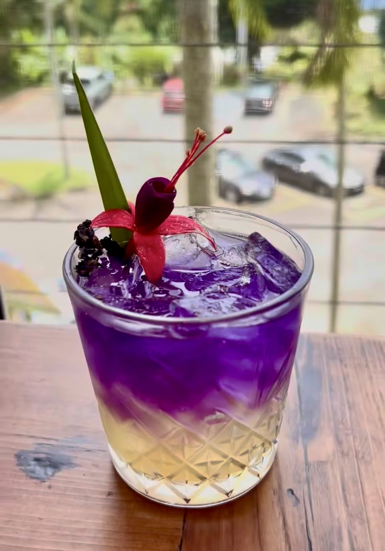 A layered purple cocktail garnished with a fuchsia flower on a wood table in the SoKo sunroom, overlooking Kainaliu